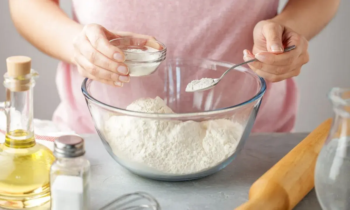 Une personne ajoute une poudre blanche dans un bol de farine, une alternative pour remplacer la levure boulangère. Huile, sel et rouleau à pâtisserie à proximité.