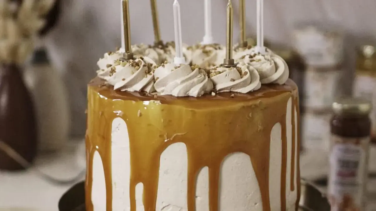 Gâteau d'anniversaire à étages, nappé de caramel beurre salé, décoré de crème et de noisettes concassées, avec des bougies dorées et blanches.
