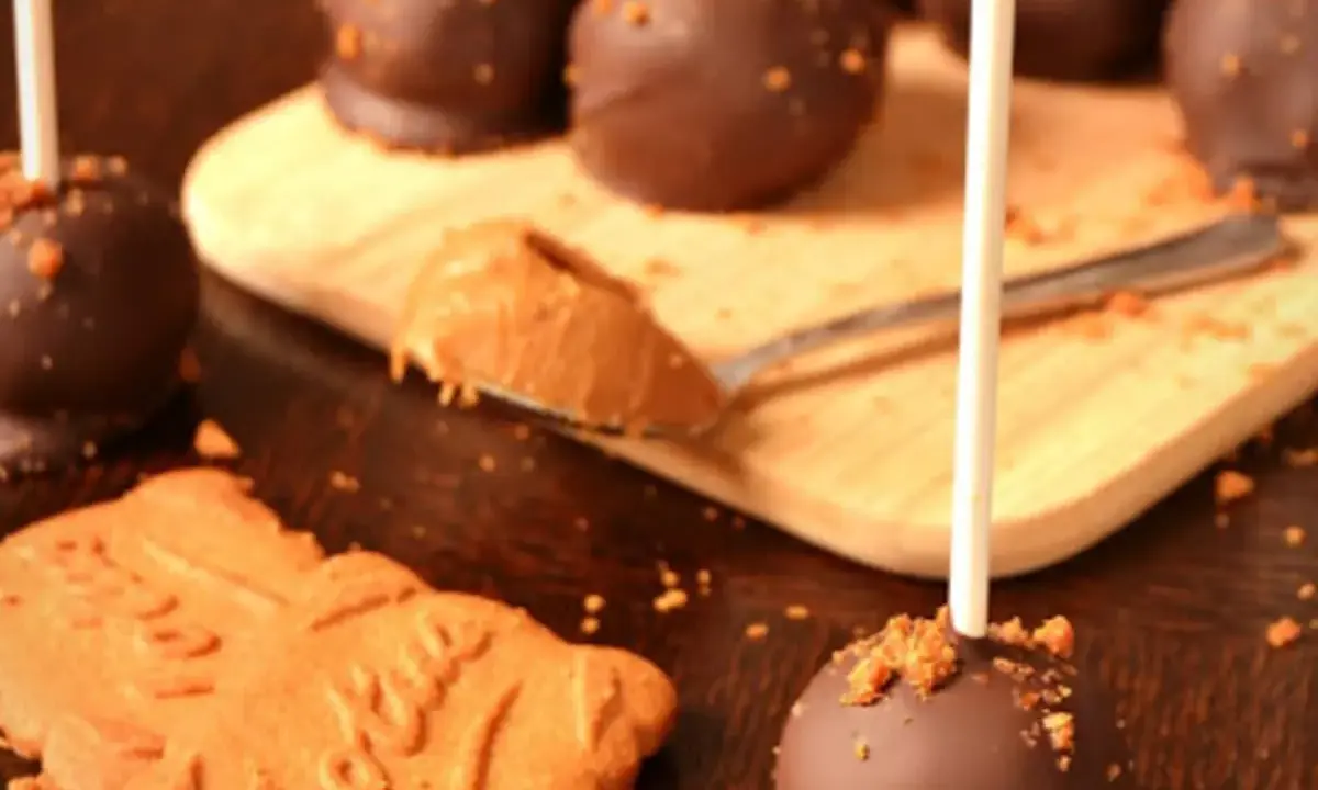 Cake pops speculoos enrobés de chocolat, parsemés de miettes de biscuit. Une cuillère de pâte à tartiner speculoos est à côté.