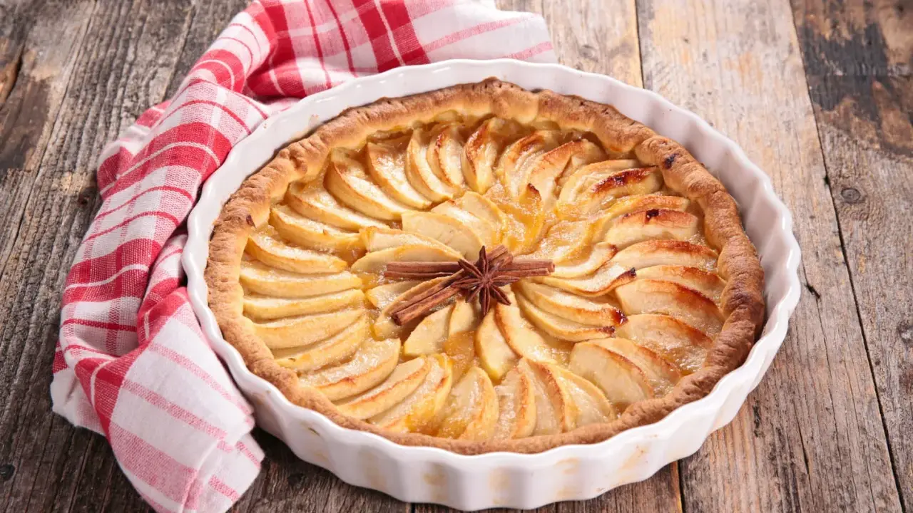 Tarte aux pommes dorée, décorée de bâtons de cannelle et d'anis étoilé. Une serviette à carreaux rouges et blancs est posée à côté. Idéale pour la conservation.