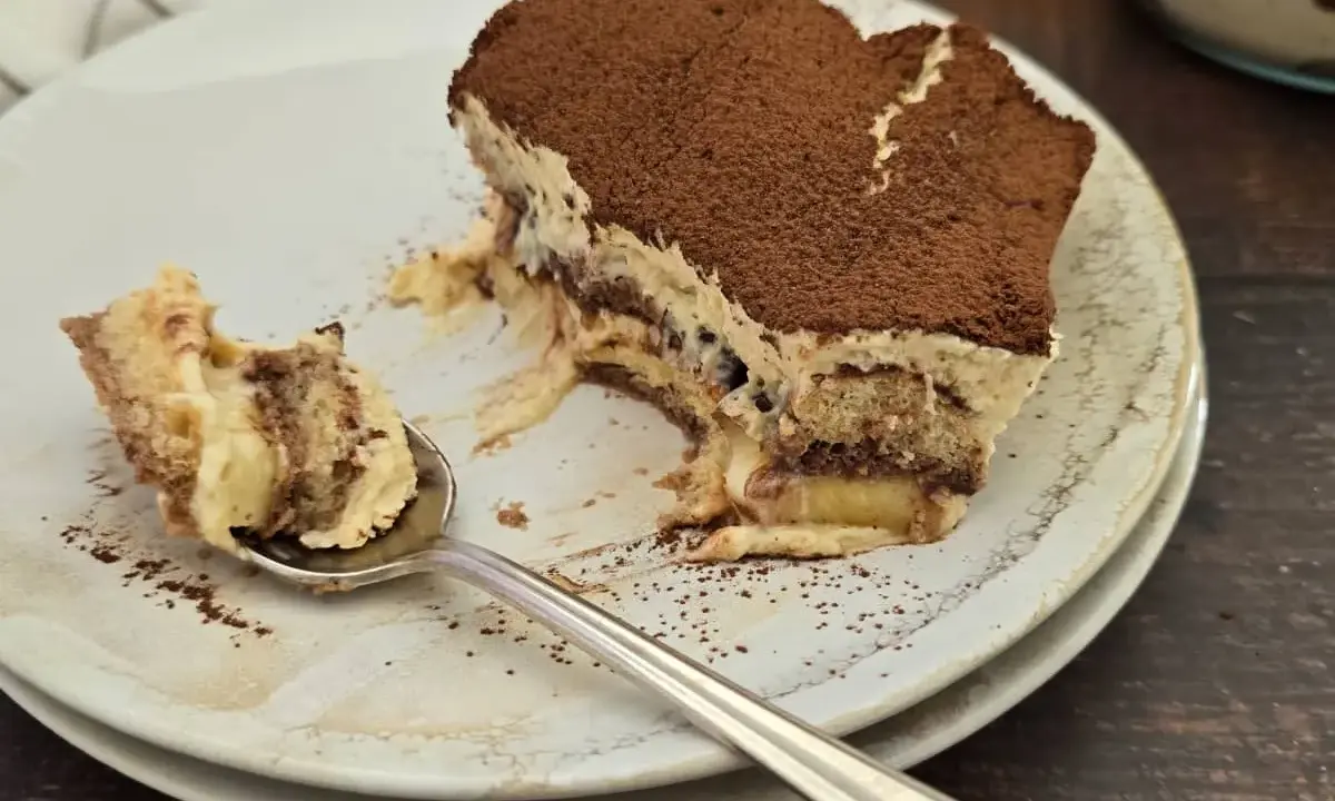Une part de tiramisu nutella, saupoudrée de cacao, sur une assiette. Une cuillère en argent a prélevé un morceau crémeux.