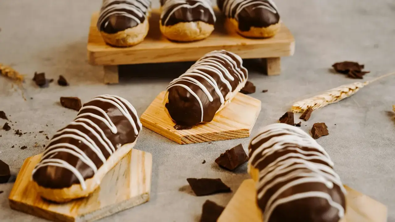 Éclairs gourmands avec un glaçage éclair chocolat brillant, décorés de fines rayures blanches. Quelques morceaux de chocolat et une tige de blé complètent la scène.
