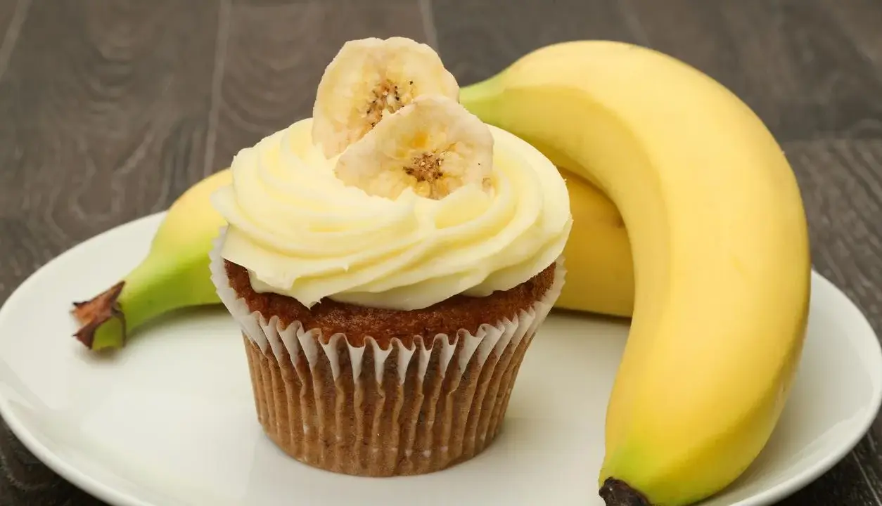Un délicieux cupcake banane, orné de fines tranches de banane séchée, repose sur une assiette blanche, à côté de bananes fraîches.