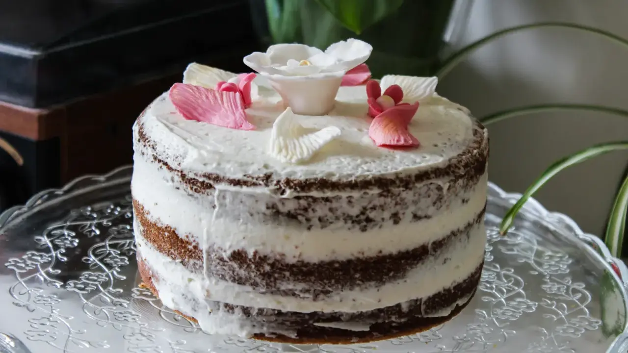 Un délicieux layer cake fleuri, avec des pétales de roses en sucre roses et blancs, posé sur un plat en verre orné.