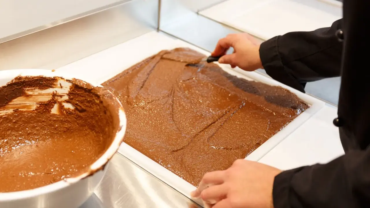 Un chef étale une pâte chocolatée onctueuse dans un moule. C'est la base du praliné, une gourmandise qui fait rêver.