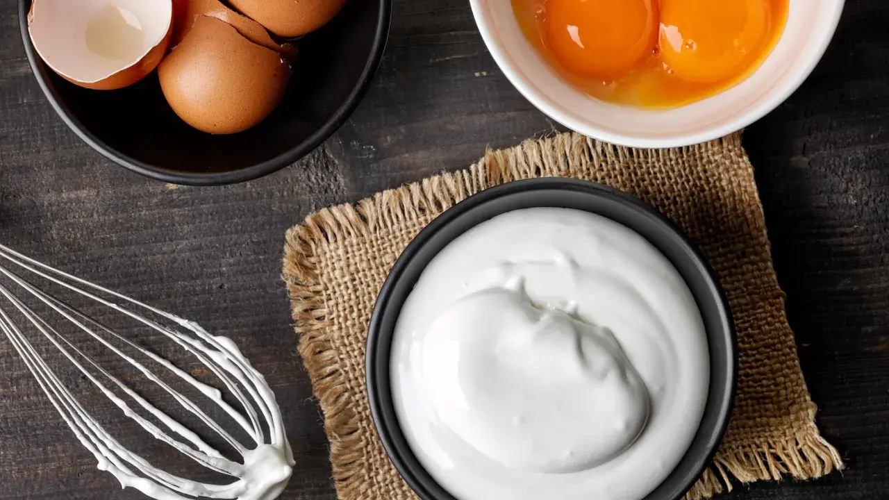 Préparation d'une meringue : blancs en neige, jaunes d'œufs, coquilles et fouet sur fond de bois sombre.