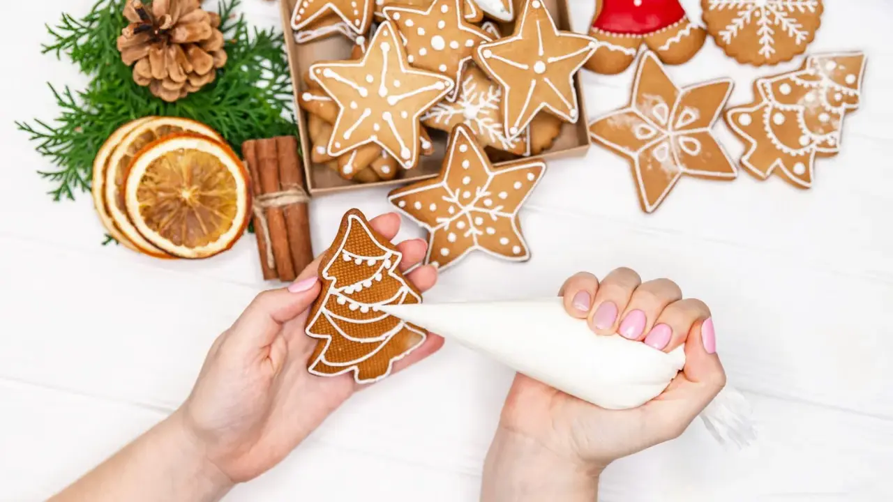 Mains décorant un biscuit sapin avec du glaçage royal. Autour, des étoiles et des décorations de Noël.
