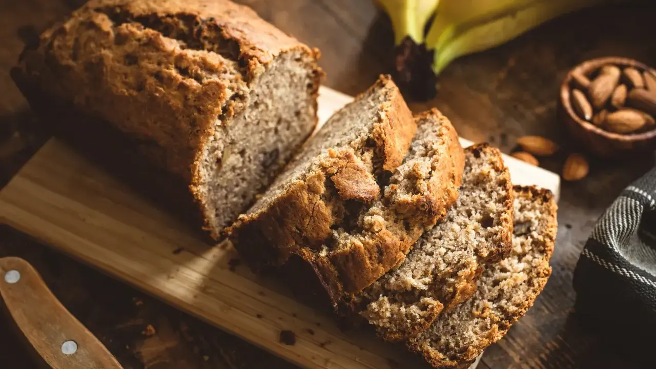 Tranches de banana bread moelleux sur une planche en bois, avec des bananes et des amandes en arrière-plan.