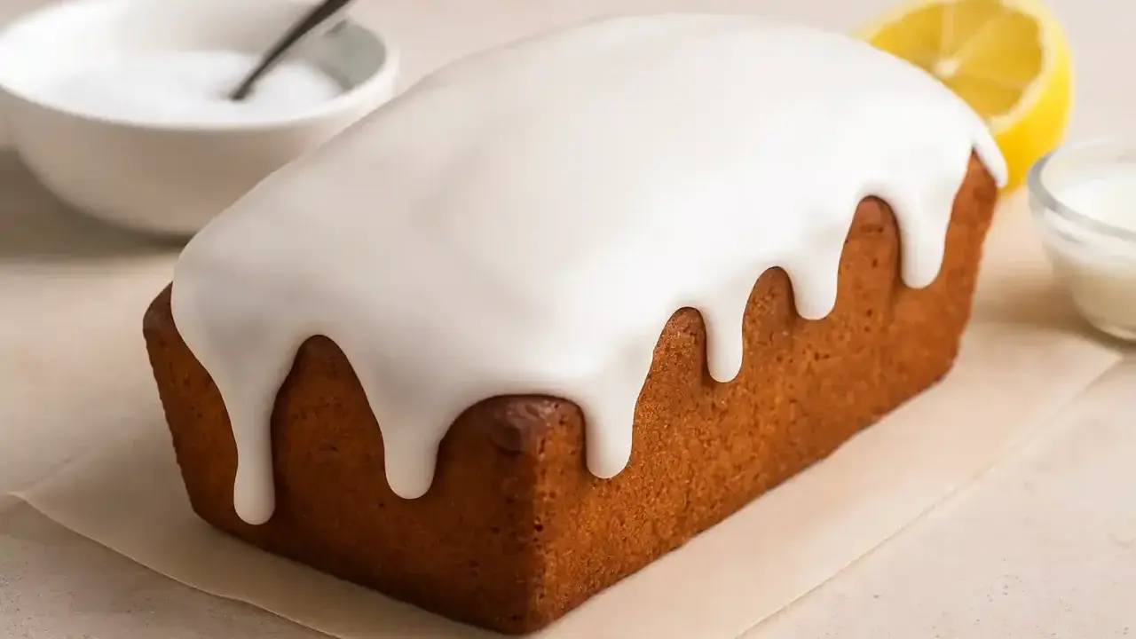 Un délicieux cake au citron recouvert d'un glaçage sucre glace facile, avec une tranche de citron et un bol d'ingrédients à proximité.