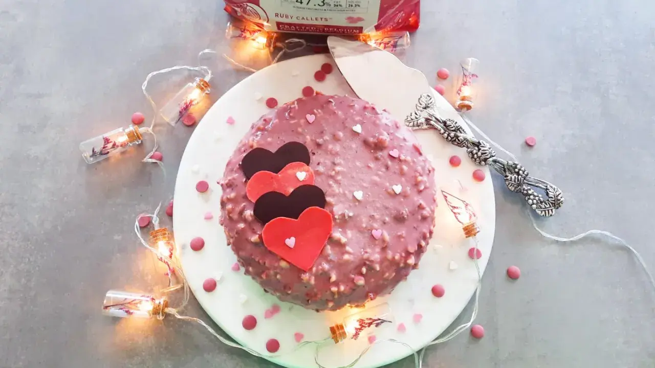 Gâteau rose orné de cœurs, enrobé de chocolat ruby croquant. Guirlande lumineuse et confettis ajoutent une touche festive.