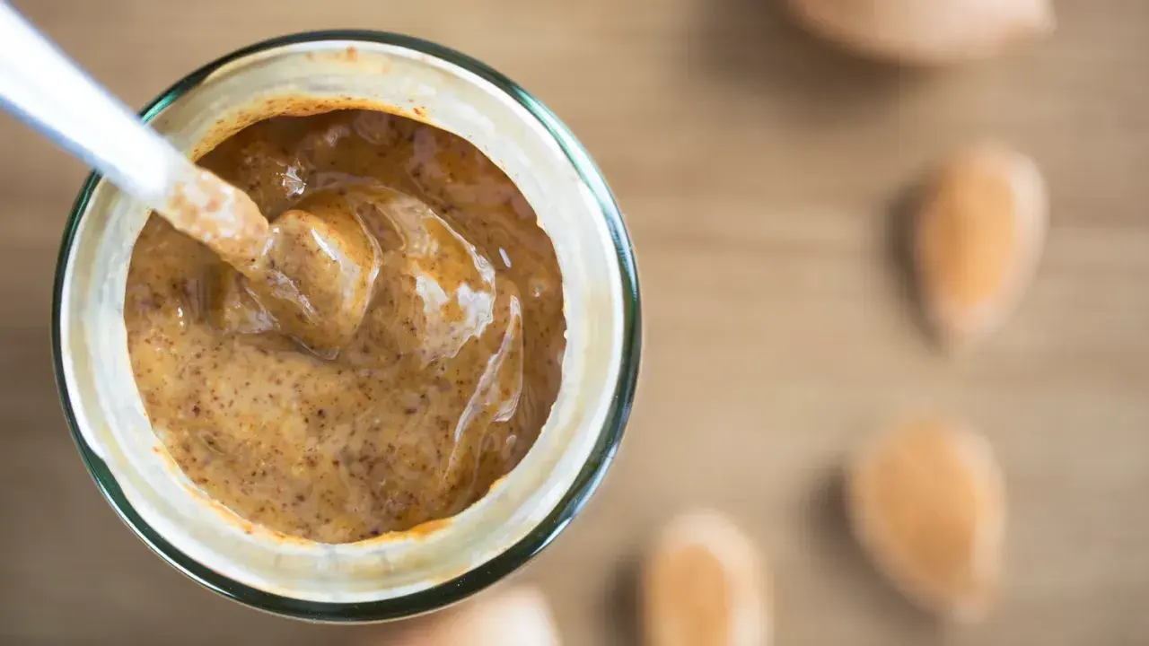 Une cuillère plonge dans un pot de purée d'amande maison, crémeuse et onctueuse. Des amandes sont disposées autour.