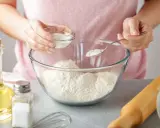 Une personne ajoute une poudre blanche dans un bol de farine, une alternative pour remplacer la levure boulangère. Huile, sel et rouleau à pâtisserie à proximité.