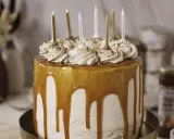 Gâteau d'anniversaire à étages, nappé de caramel beurre salé, décoré de crème et de noisettes concassées, avec des bougies dorées et blanches.