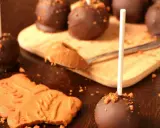Cake pops speculoos enrobés de chocolat, parsemés de miettes de biscuit. Une cuillère de pâte à tartiner speculoos est à côté.