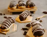 Éclairs gourmands avec un glaçage éclair chocolat brillant, décorés de fines rayures blanches. Quelques morceaux de chocolat et une tige de blé complètent la scène.