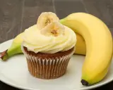 Un délicieux cupcake banane, orné de fines tranches de banane séchée, repose sur une assiette blanche, à côté de bananes fraîches.