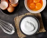 Préparation d'une meringue : blancs en neige, jaunes d'œufs, coquilles et fouet sur fond de bois sombre.