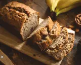 Tranches de banana bread moelleux sur une planche en bois, avec des bananes et des amandes en arrière-plan.