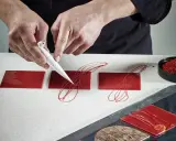 Pâtissier décorant des carrés de chocolat rouge avec un cornet. Le papier guitare, c'est quoi ? C'est ce support qui permet ces décorations fines.