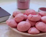 Un bol de coques macarons roses craquelées, prêtes pour la conservation. Un livre et un pot de confiture rose en arrière-plan.