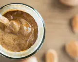 Une cuillère plonge dans un pot de purée d'amande maison, crémeuse et onctueuse. Des amandes sont disposées autour.