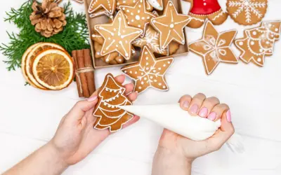 Mains décorant un biscuit sapin avec du glaçage royal. Autour, des étoiles et des décorations de Noël.