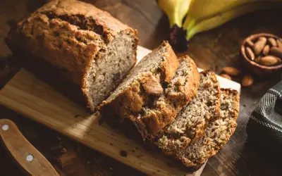 Tranches de banana bread moelleux sur une planche en bois, avec des bananes et des amandes en arrière-plan.
