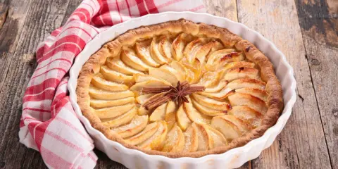 Tarte aux pommes dorée, décorée de bâtons de cannelle et d'anis étoilé. Une serviette à carreaux rouges et blancs est posée à côté. Idéale pour la conservation.