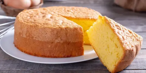 Un moelleux gâteau de Savoie, parfait pour la conservation, avec une part coupée. Des œufs et un fouet en arrière-plan suggèrent sa préparation.