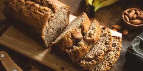 Tranches de banana bread moelleux sur une planche en bois, avec des bananes et des amandes en arrière-plan.