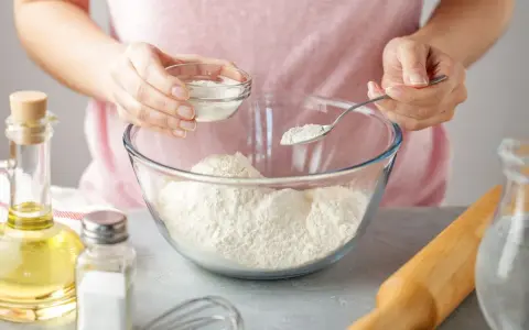 Une personne ajoute une poudre blanche dans un bol de farine, une alternative pour remplacer la levure boulangère. Huile, sel et rouleau à pâtisserie à proximité.