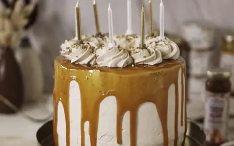 Gâteau d'anniversaire à étages, nappé de caramel beurre salé, décoré de crème et de noisettes concassées, avec des bougies dorées et blanches.