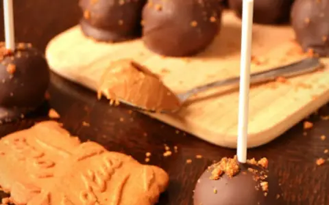 Cake pops speculoos enrobés de chocolat, parsemés de miettes de biscuit. Une cuillère de pâte à tartiner speculoos est à côté.
