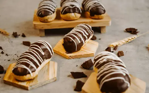 Éclairs gourmands avec un glaçage éclair chocolat brillant, décorés de fines rayures blanches. Quelques morceaux de chocolat et une tige de blé complètent la scène.