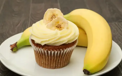 Un délicieux cupcake banane, orné de fines tranches de banane séchée, repose sur une assiette blanche, à côté de bananes fraîches.