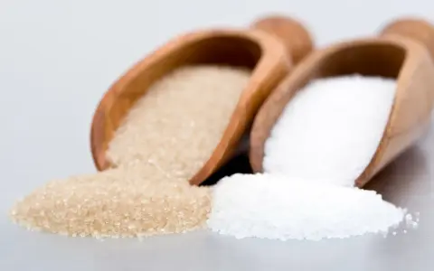 Deux cuillères en bois contiennent différents types de sucre en pâtisserie : du sucre blanc et du sucre roux.