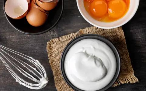 Préparation d'une meringue : blancs en neige, jaunes d'œufs, coquilles et fouet sur fond de bois sombre.
