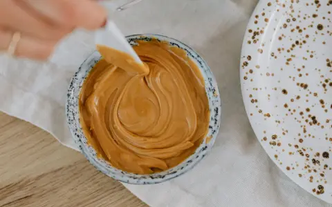 Une spatule prélève une portion de crème d'amandes légère dans un bol. Une assiette tachetée est à côté.