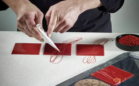 Pâtissier décorant des carrés de chocolat rouge avec un cornet. Le papier guitare, c'est quoi ? C'est ce support qui permet ces décorations fines.