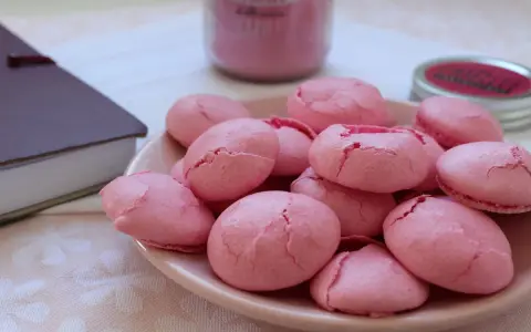 Un bol de coques macarons roses craquelées, prêtes pour la conservation. Un livre et un pot de confiture rose en arrière-plan.