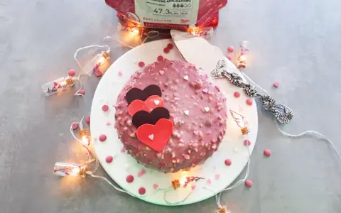Gâteau rose orné de cœurs, enrobé de chocolat ruby croquant. Guirlande lumineuse et confettis ajoutent une touche festive.