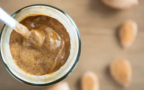 Une cuillère plonge dans un pot de purée d'amande maison, crémeuse et onctueuse. Des amandes sont disposées autour.