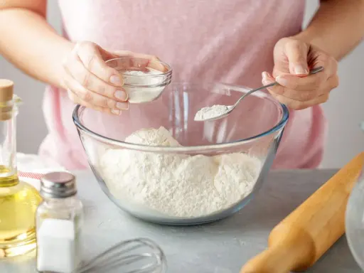 Une personne ajoute une poudre blanche dans un bol de farine, une alternative pour remplacer la levure boulangère. Huile, sel et rouleau à pâtisserie à proximité.