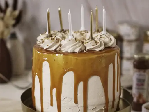 Gâteau d'anniversaire à étages, nappé de caramel beurre salé, décoré de crème et de noisettes concassées, avec des bougies dorées et blanches.