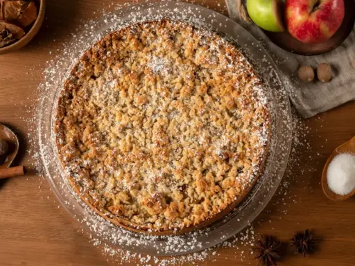 Un délicieux crumble pomme, saupoudré de sucre glace, entouré de pommes fraîches, de noix de muscade et de bâtons de cannelle.