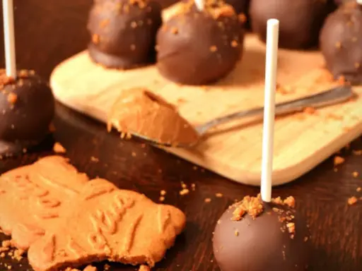 Cake pops speculoos enrobés de chocolat, parsemés de miettes de biscuit. Une cuillère de pâte à tartiner speculoos est à côté.
