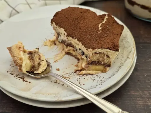 Une part de tiramisu nutella, saupoudrée de cacao, sur une assiette. Une cuillère en argent a prélevé un morceau crémeux.