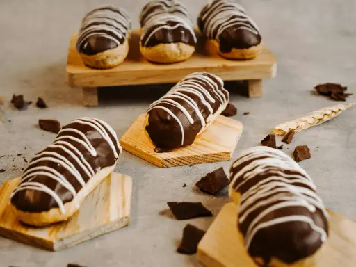 Éclairs gourmands avec un glaçage éclair chocolat brillant, décorés de fines rayures blanches. Quelques morceaux de chocolat et une tige de blé complètent la scène.