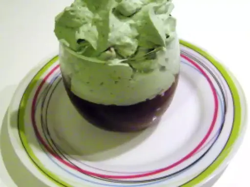 Un verre de dessert avec une couche de chocolat au fond et une généreuse chantilly pistache aérienne sur le dessus, posé sur une assiette colorée.
