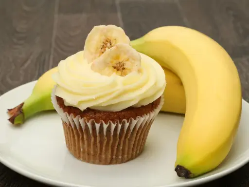 Un délicieux cupcake banane, orné de fines tranches de banane séchée, repose sur une assiette blanche, à côté de bananes fraîches.